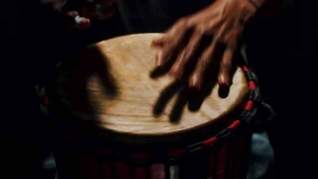 Black musician playing the wooden hand drum close up in a dark studio. Music performer vigorously beating the traditional djembe drum with hands