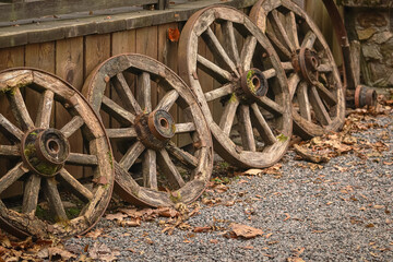 Fototapeta na wymiar Old wooden wheels arranged in a row near wooden plank elevation on pebble road with autumn leaves