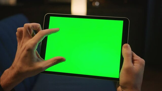 Close-up footage of unrecognizable man sitting on sofa and using blank large green screen tablet in evening at home. Fingers pinching to zoom tablet's screen. Indoors