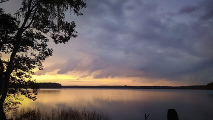 Sunset sky over lake landscape, nature background with heavy clouds and tree silhouette
