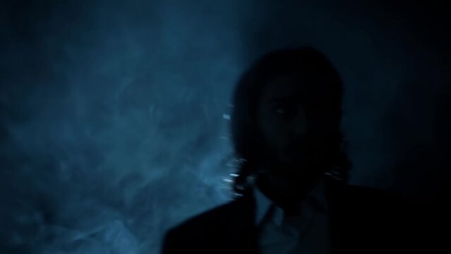 Cinematic shot of a young man in a blue suit with long hair and blood on his face, coming upstairs looking sideways, and keeping a gun on his shoulder. HD footage with smoke element 24FPS.
