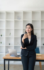 Portrait of young confident woman holding coffee cup, standing at office and looking outside. Successful businesswoman standing in office with copy space.