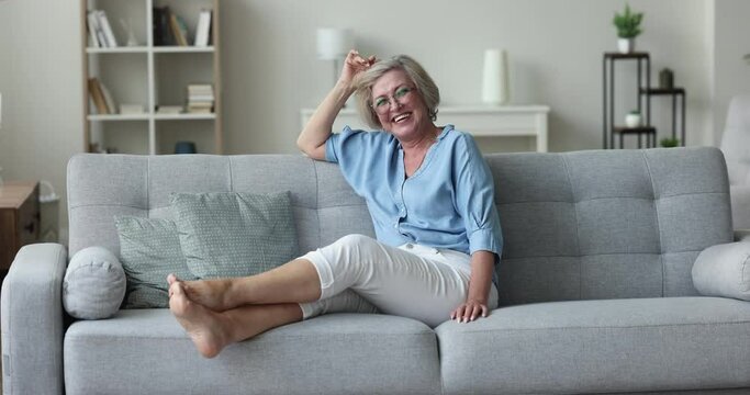 Overjoyed mature woman relaxing on cozy sofa in warm living room laughing smile staring at camera looks carefree feels happy to spend leisure at own modern flat, having untroubled life on retirement
