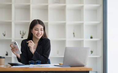 Smiling beautiful Asian businesswoman analyzing chart and graph showing changes on the market and holding smartphone at office.