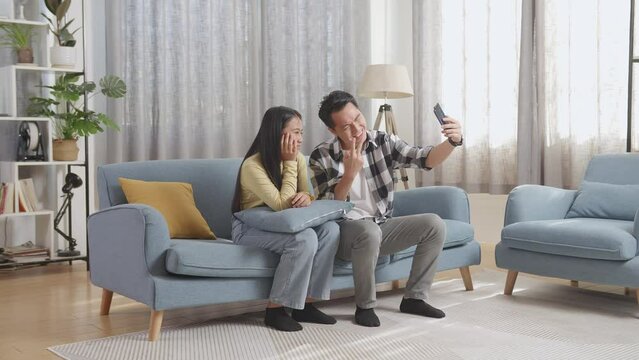 Smiling Asian Father And His Daughter Showing Peace And Thumbs Up Gestures While Taking A Picture On A Sofa In The Living Room At Home
