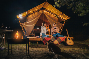 Fototapeta na wymiar Two Smiling female friends drinking wine and eating fruits sitting in cozy glamping tent in autumn evening bonfire. Luxury camping tent for outdoor holiday and vacation. Lifestyle concept