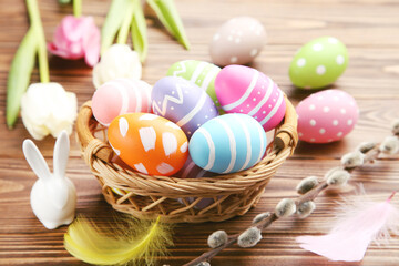 Fototapeta na wymiar Colorful eggs in basket and porcelain rabbit figurine, feathers, flowers of tulips and branches of willow on brown wooden background