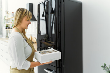 A woman pastry chef in the kitchen in an apron stands near a black refrigerator and holds a box of ready-made sweets in her hand for storage in the cold