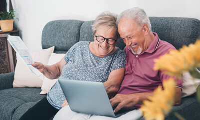 Aged couple at home paying bills on line with laptop and laughing a lot having fun together. Happiness and elderly lifestyle. Man and woman old senior sitting on sofa with computer online connection - obrazy, fototapety, plakaty