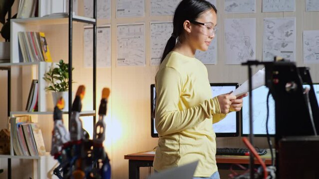 Teen Asian Girl Looking At The Picture In Hand, Thinking, And Walking Around While Working About A Cyborg Hand At Home
