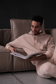 Portrait of a cheerful young african american man 20s wearing casual hoody and glasses sitting on the floar near the sofa against black background with magazine 