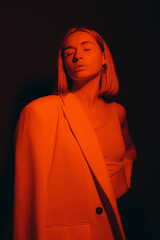 Serene female model in trendy jacket closing eyes while standing against black backdrop in dark studio