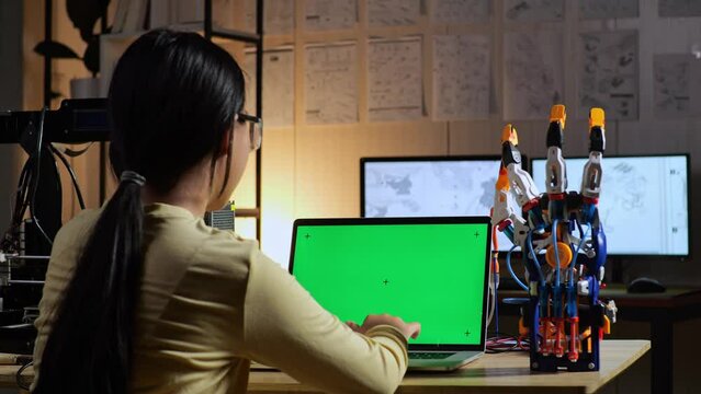 Back View Of Teen Asian Girl Designing A Cyborg Hand On A Laptop With Mock Up Green Screen At Home

