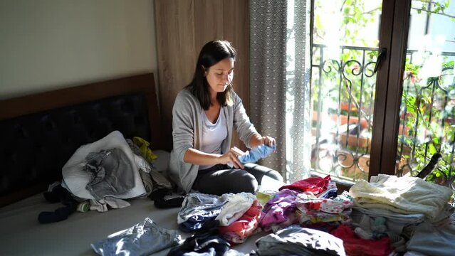 Mom Stacks Clean Baby Clothes In Piles While Sitting On The Bed Next To A Playing Child