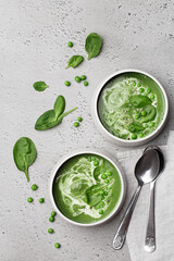 Obraz premium Two ceramic bowls with spinach cream soup with green pea and fresh spinach leaves on grey concrete background. Top view