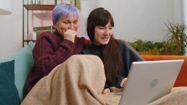 Two lesbian women couple or girls friends looking at laptop camera, making video webcam conference call with family, enjoying pleasant conversation, laughing, waving hello at home room. LGBT people