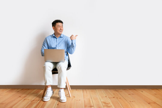 Excited Middle Aged Asian Man Sitting On Chair With Laptop And Pointing Aside At Free Space Over White Studio Wall