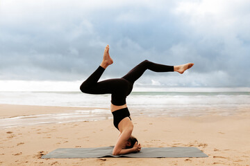 Young woman practicing yoga on the beach by seaside, doing headstand exercise on fitness mat, side view, copy space