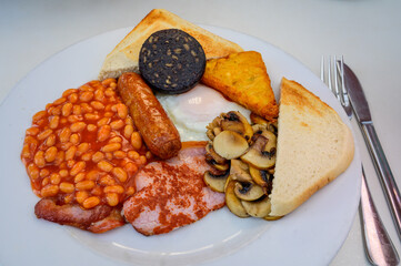 White board with full English breakfast with bacon, fried egg, beans, tomato, roasted sausage, black pudding, scons, hash browns and fried mushrooms