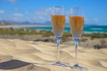 Two glasses of rose champagne or cava sparkling wine served on white sandy tropical beach with dunes and blue ocean, romantic vacation, winter sun on Fuerteventura, Canary, Spain