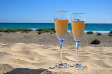 Two glasses of rose champagne or cava sparkling wine served on white sandy tropical beach with dunes and blue ocean, romantic vacation, winter sun on Fuerteventura, Canary, Spain