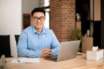 Portrait of happy middle aged asian businessman sitting at desk with laptop in home office and...