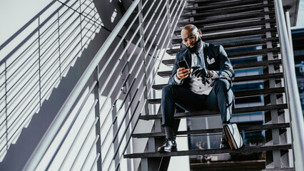 An elegant bald black businessman wearing glasses and a tailored suit, and black leather gloves, is sitting on a step of an outdoor stairway with a briefcase by his foot while on his phone texting