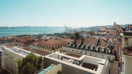 A sunny shot of Lisbon city center, a cluster of restored buildings, with red brick roofing, for housing, and local businesses, and in the background the 25th of April Bridge can be seen far away