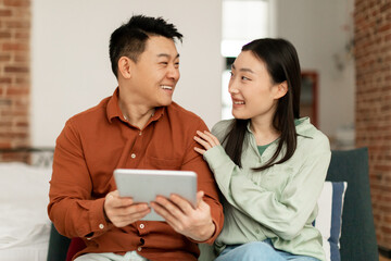 Happy asian middle aged man and young woman sitting on sofa and using digital tablet, looking at each other and smiling
