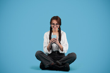 Smiling caucasian teen girl with pigtails student sit on floor, chatting on phone, isolated on blue background