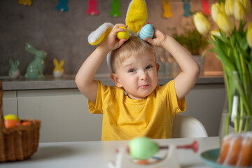 Emotional portrait of a cheerful little boy wearing bunny ears on Easter day who laughs merrily, plays with colorful Easter eggs sitting at a table in the kitchen.