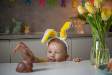 A cute little boy wearing bunny ears on Easter day wants to eat a chocolate easter bunny. A child...