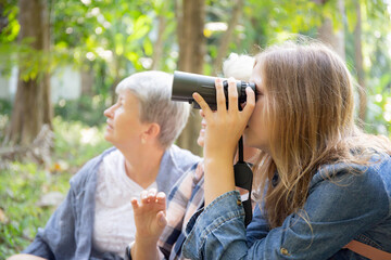 Teenager girl and two grandmother traveling travel to forest and adventure while using binoculars exploration together with happy, senior woman and granddaughter hiking with fun and vitality.