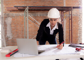 Young caucasian architect woman looking laptop and working drawing blueprint for planning building house on desk, engineer thinking and sketching blueprint for construction, industrial concept.