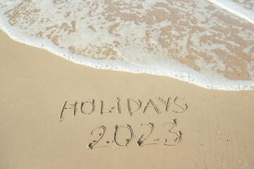 Holidays 2023 lettering on the beach with wave and clear blue sea. 