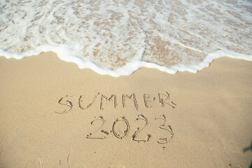 Summer 2023 lettering on the beach with wave and clear blue sea. 
