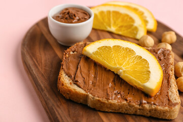 Wooden board of tasty toast with hazelnut butter and orange slices on pink background, closeup