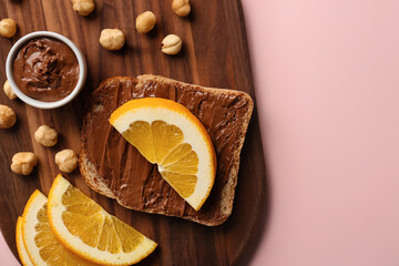 Wooden board of tasty toast with hazelnut butter and orange slices on pink background, closeup