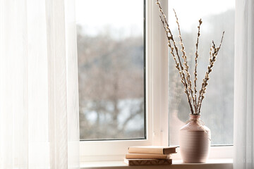 Vase with pussy willow branches and books on windowsill in room