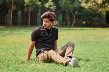 Attractive young indian man portrait in black t shirt and silver neck chain sitting on green lawn...