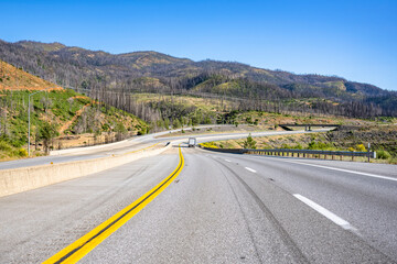 Big rig semi truck transporting cargo in dry van semi trailer running downhill on the winding highway road between the hills