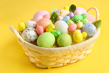 Basket with beautiful Easter eggs and plant leaves on yellow background