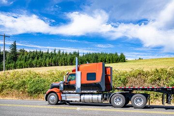 Fototapeta na wymiar Stylish black and orange big rig semi truck transporting empty flat bed semi trailer driving on the road along the summer meadow with green grass and line of trees