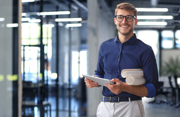 Confident young business man in shirt examining blueprint while standing against a window at office.