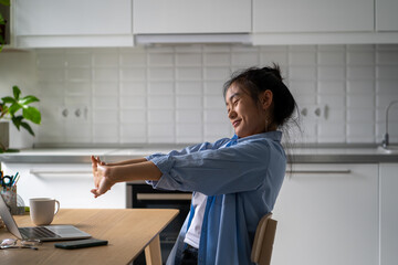 Sleepy young Asian woman freelancer sitting at desk with laptop computer stretching arms, preparing to work remotely. Smiling relaxed girl remote worker resting at home office. Freelance productivity