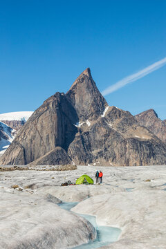 Two mountaineers stand next to tent on glacier under mountain summit.