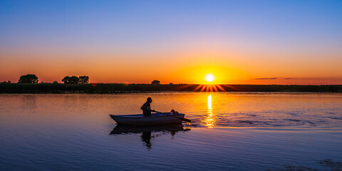 A classic rowboat against a backlit orange sunset in semi-darkness. Silhouette of a 60-year-old...