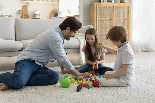 Happy daddy engaged in game with cheerful little son and daughter kids, playing toy blocks on soft carpeted floor, arranging cubes from heap, talking, smiling, laughing, enjoying leisure, playtime
