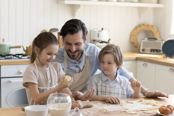 Happy positive dad and little son and daughter kids in aprons baking in kitchen, shaping dough at floury kitchen table, preparing bakery food, dessert, cooking together, talking, smiling