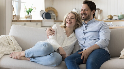 Obraz premium Happy cheerful dating couple in love enjoying leisure at home together, hugging on soft couch, looking away, smiling, laughing, watching movie, TV, discussing plans. Banner shot
